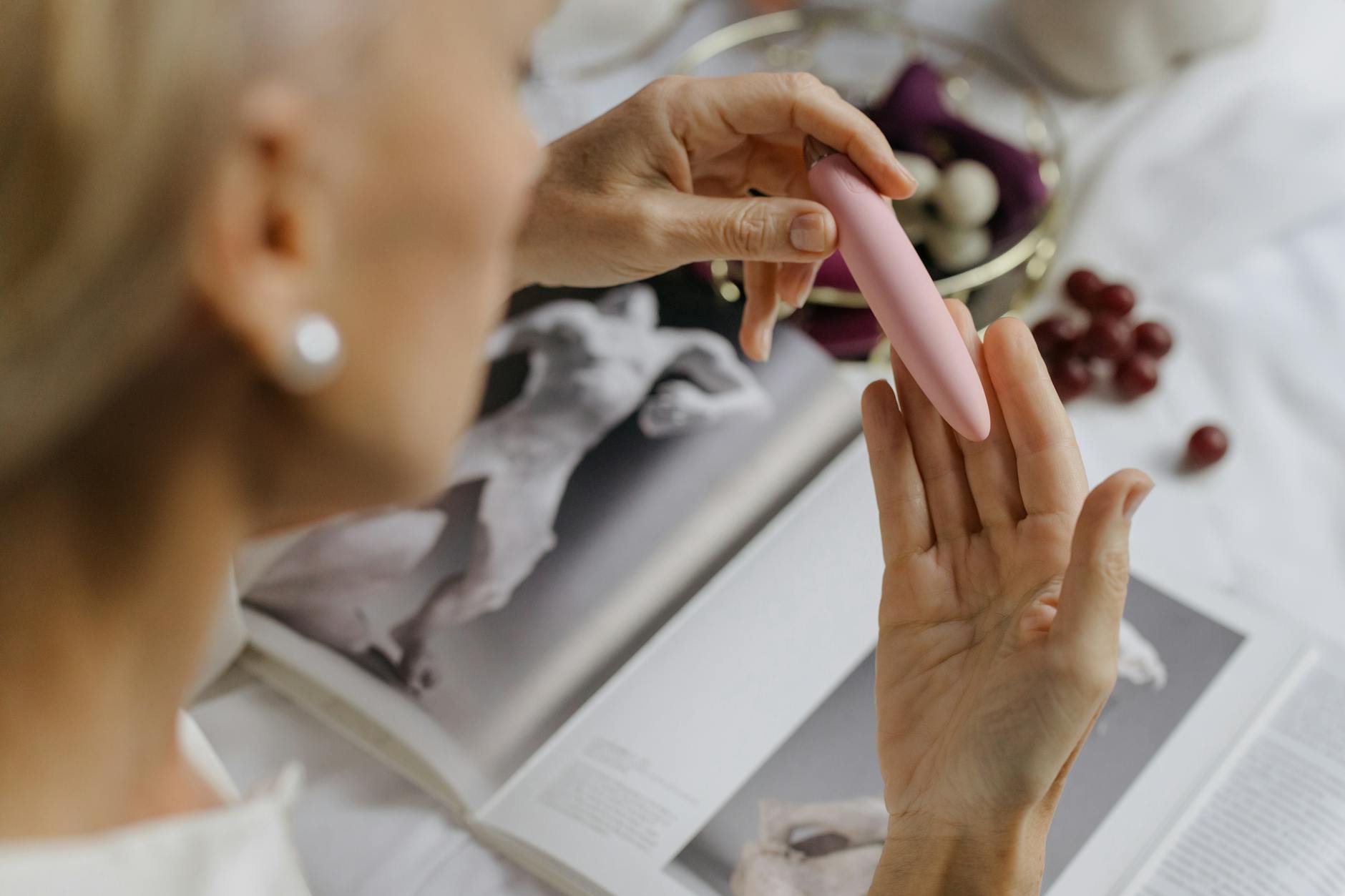 Close-up of a hand holding a pink lemon vibrator, with art books in the background, emphasizing intimate and personal exploration.