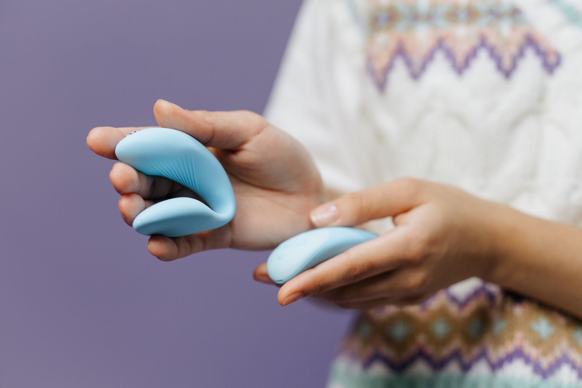 Close-up of hands holding a sleek clitoral vibrator against a purple background, showing intimate connection to pleasure devices.